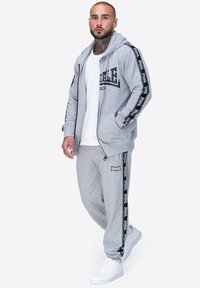 Sudadera con cremallera gris con detalles en negro, combinada con pantalones de chándal a juego que presentan rayas laterales. Zapatillas blancas completan el conjunto.