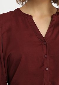 Blusa bordeaux con scollatura a V, chiusura con bottoni e maniche corte. Realizzata in un tessuto leggero e liscio con una leggera lucentezza.