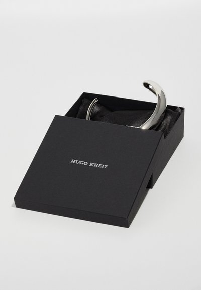 Zilveren gebogen accessoire in een zwarte doos met een zachte stoffen voering, featuring het label "HUGO KREIT" in opvallende zilveren letters op de deksel.