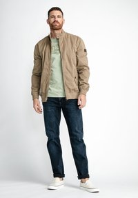 Chaqueta bomber marrón claro, camiseta verde, jeans de mezclilla oscuros y zapatos slip-on blancos; presenta un cuello alto y cierre frontal.