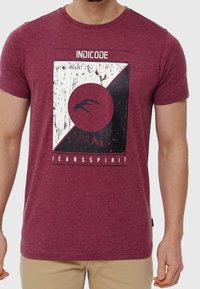 T-shirt in cotone bordeaux con un design grafico che presenta accenti bianchi e neri, inclusi un motivo circolare e il testo "INDICODE JEANSSPIRIT."