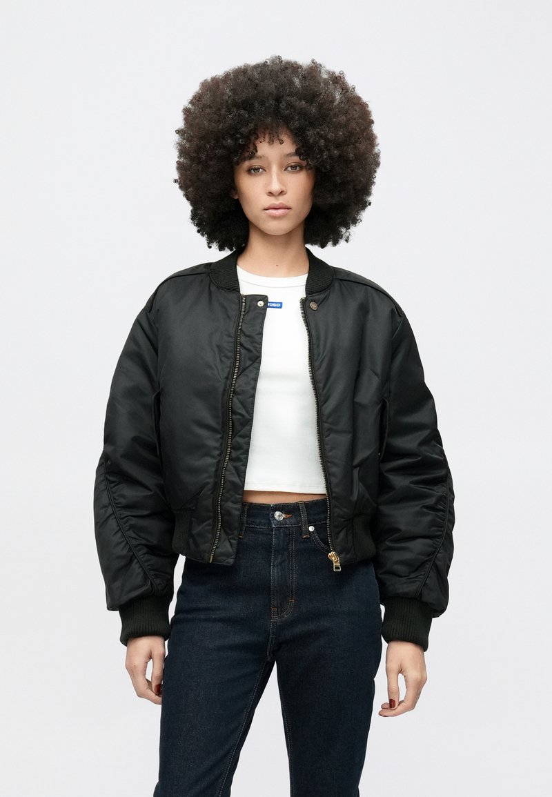 Femeie cu păr creț și voluminos, purtând o jachetă bomber neagră peste un tricou alb scurt și blugi înalți pe talie de culoare închisă, stând în picioare pe un fundal simplu.