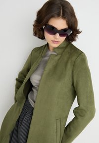 Veste en daim vert avec un col cranté, poches latérales, associée à un haut gris et à un pantalon rayé. Le mannequin porte des lunettes de soleil noires élégantes.
