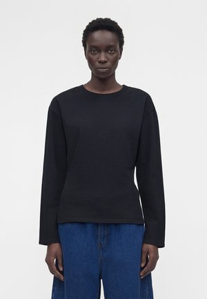 Personne avec des cheveux courts et bouclés portant un haut noir uni à manches longues et un jean bleu, debout devant un fond blanc.