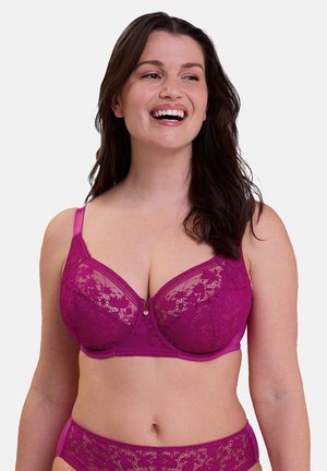 Lachende vrouw die een bijpassende paarse kanten bh en ondergoedset draagt, lichtjes opzij kijkend tegen een effen achtergrond.