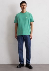 T-shirt in cotone verde menta con un piccolo logo bianco sul petto, abbinato a pantaloni in denim blu scuro e scarpe nere stringate.