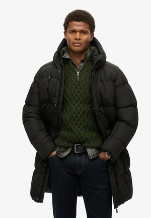 Parka inflable en negro, con capucha y diseño acolchado. Debajo, se puede ver un suéter de punto de cable en verde oscuro y una camisa de cuadros.