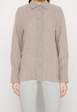 Nainen, jolla on pitkähihaiset beige pellavapaita, jossa napit edessä, ja vaaleanharmaat housut yksiväristä taustaa vasten.