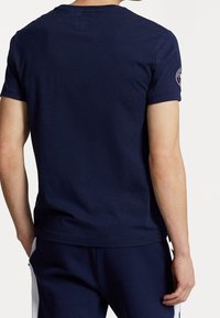 Muž stojící zády k fotoaparátu, oblečený v tmavě modrém tričku s krátkým rukávem a odpovídajících tmavě modrých kalhotách s bílými postranními pruhy.