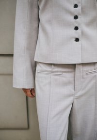 Világosszürke, testre szabott öltöny fekete gombokkal a zakón, semleges háttér előtt a törzset és egy kart mutatva.