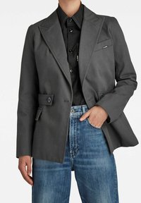 Harmaa raidallinen bleiseri, jossa on säädetty istuvuus, vekatut kaulukset ja kaksi etutaskua, yhdistetty mustan paidan ja sinisten farkkujen kanssa.