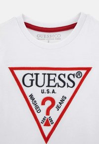 Bílý organický bavlněný tričko s výšivkou červeného a černého trojúhelníkového loga s nápisy "GUESS", "WASHED" a žerzejovými detaily na výstřihu.