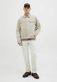 Giacca di jeans beige con due tasche sul petto, indossata sopra un top grigio. Il modello indossa pantaloni bianchi e scarpe marroni, su uno sfondo chiaro.