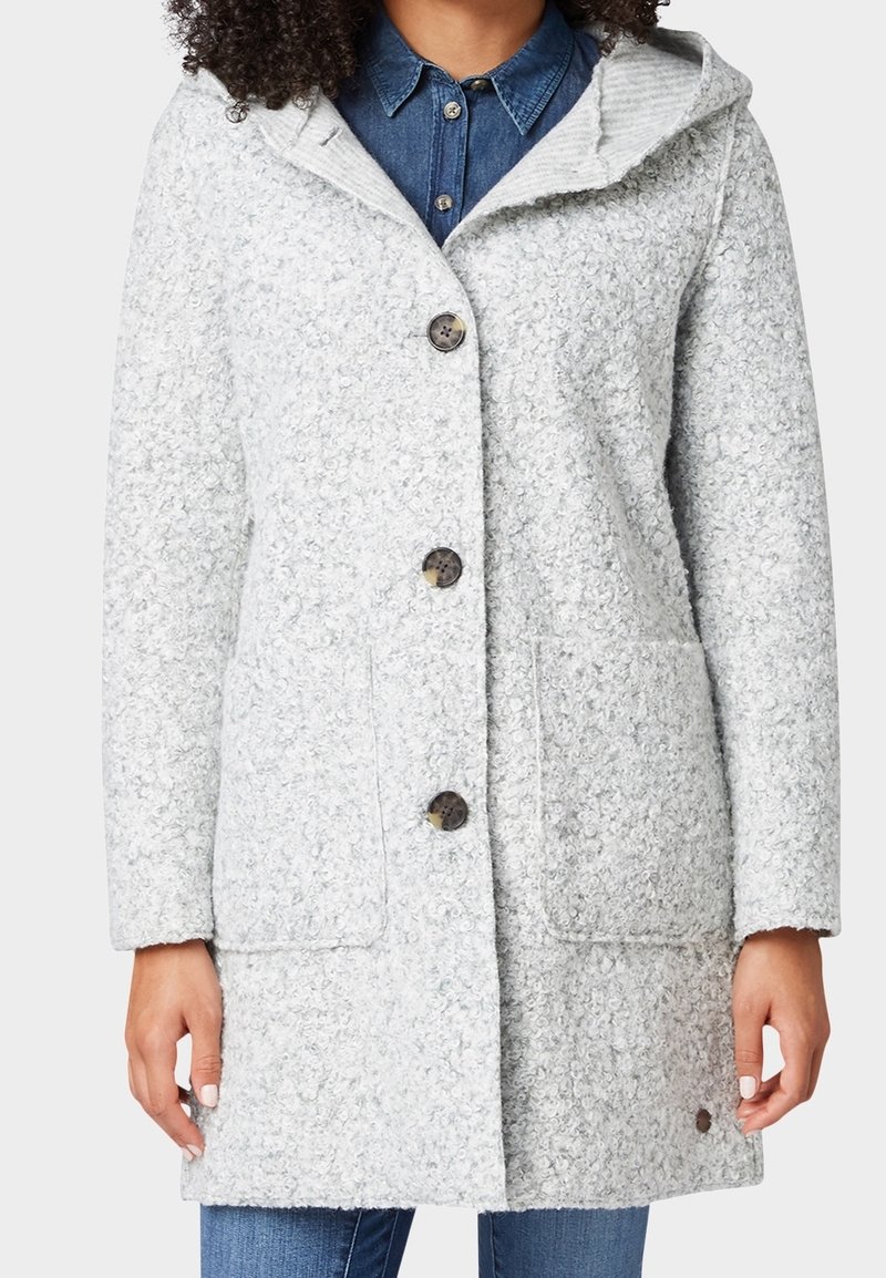 Šedý texturovaný kabát s kapucí, který má velké knoflíky, dvě přední kapsy a pletený detail na límci. Vzhled je po kolena.