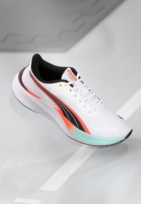 Weißer Sportschuh mit schwarzen und korallenfarbenen Akzenten, strukturiertes Mesh-Obermaterial, gepolsterte Sohle und auffälligem Markenlogo auf der Zunge.