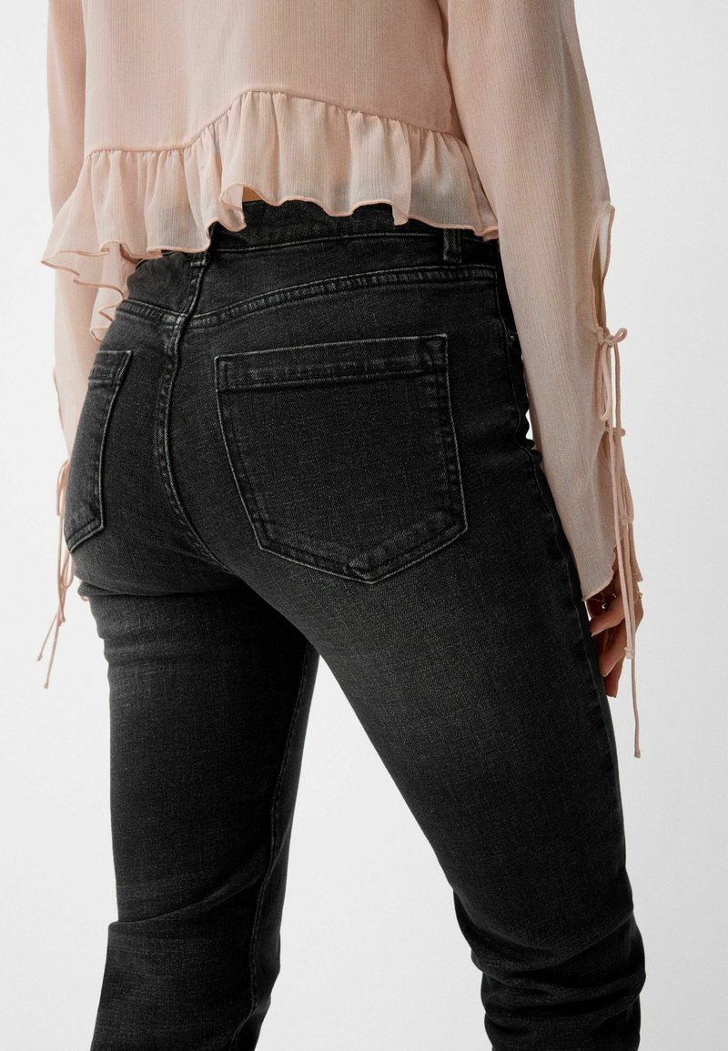 Jeans in denim nero con vestibilità slim, dotati di tasche posteriori. Abbinati a una blusa rosa chiaro con orlo arricciato e lacci ai polsini delle maniche.
