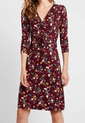 Femme portant une robe bordeaux mi-longue avec manches trois-quarts et motif floral multicolore, avec un décolleté en V et une ceinture à la taille.