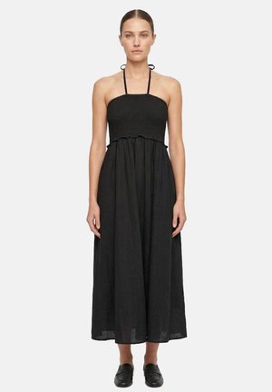 Donna che indossa un abito midi nero senza maniche con scollo halter e gonna arricciata e scarpe nere slip-on, in piedi davanti a uno sfondo semplice.