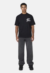 T-shirt in cotone nero con la scritta "Good for Nothing" in bianco sul davanti, abbinata a jeans neri sbiaditi e larghi e sneakers nere.