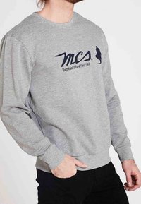 Homme portant un sweat-shirt gris avec un texte noir « MCS Rugged and Refined Since 1987 » et un petit emblème en silhouette sur la poitrine.