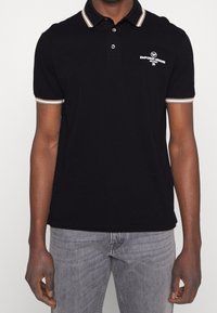 Man i svart pikétröja med beige-randig krage och ärmar, prydd med vit Emporio Armani Milano-logotyp på bröstet, tillsammans med grå jeans.