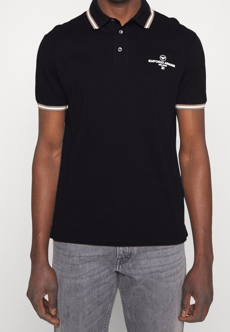 Man i svart pikétröja med beige-randig krage och ärmar, prydd med vit Emporio Armani Milano-logotyp på bröstet, tillsammans med grå jeans.