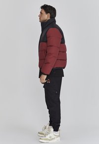 Chaqueta de plumas roja y negra con un alto cuello, con gruesos pliegues horizontales. Llevada sobre pantalones cargo negros y zapatillas en tonos neutros.