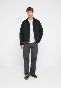 Black zipped jacket with a collar, gray sweatshirt underneath, gray trousers, and black shoes with white accents. Plain background.