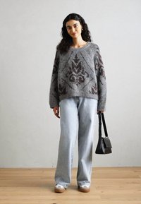 Grauer Strickpullover mit braunen geometrischen Mustern, kombiniert mit hellblauen Weitbeinen-Jeans und einer kleinen schwarzen Handtasche. Glatte Textur, lässige Passform.