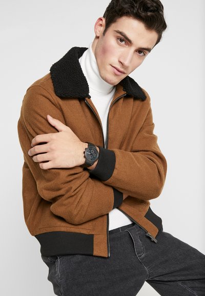 Joven con chaqueta marrón con cuello y puños negros, cuello vuelto blanco, vaqueros negros y reloj de pulsera negro, posando contra un fondo claro y liso.