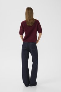 Femme avec des cheveux raides jusqu'aux épaules portant un pull court à manches courtes bordeaux et un jean droit bleu foncé, se tenant les mains sur les hanches.