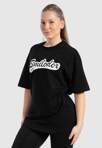 Camiseta de algodón negra con el logotipo blanco "Smilodox", mangas cortas, corte relajado y escote redondo. Costuras acentuadas en los laterales.