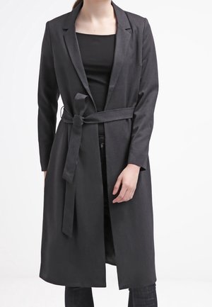 Mujer con un abrigo largo gris oscuro con cinturón sobre una camiseta negra y jeans oscuros, de pie contra un fondo blanco liso.