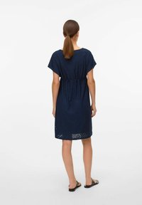 Abito blu navy con maniche corte, vestibilità comoda e motivo ad occhiello intricato; vita arricciata, arriva sopra le ginocchia, indossato con sandali bassi.