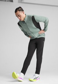 Trainingsjack in mintgroen met zwarte zijpanelen, gecombineerd met zwarte sportbroek. Heeft een lichtgewicht stof en een rits aan de voorkant.