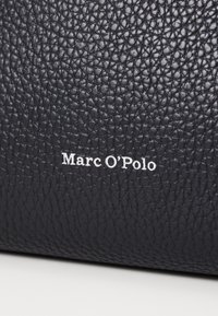 Sac à main en cuir noir avec une texture granuleuse et le logo en relief "Marc O'Polo" en blanc. Vue rapprochée mettant en valeur le matériau et l'artisanat.