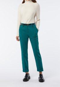 Teal korkeavyötäröiset housut, joissa on muotoiltu istuvuus, ja joissa on ruskea vyö. Yhdistettynä kermaiseen ribattuun poolopaitaan ja mustiin kenkiin.