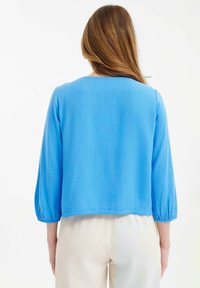 Blusa leggera blu con scollatura rotonda e maniche a tre quarti. Tessuto liscio e polsi elasticizzati. Pantaloni bianchi visibili.
