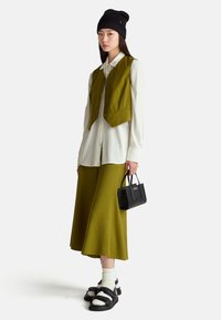Toppe in verde oliva corto sopra una camicia bianca con bottoni, abbinato a una gonna lunga verde oliva, calzini bianchi a coste, sandali neri e una borsa nera.