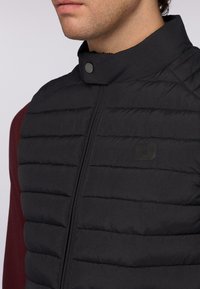 Černá prošívaná vesta má šité přihrádky, vysoký límec a přední zip. Na levé straně je viditelné malé logo.