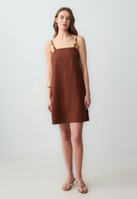 Braun Leinen ärmelloses Kleid mit dekorativen Perlenakzenten an den Trägern. A-Linien-Silhouette, minimalistisches Design und strukturierter Stoff. Sichtbares lässiges Schuhwerk.