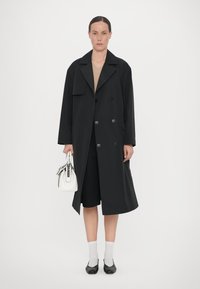 Must musta kahekordse nööbiga trench coat suurte kraedega, millele on lisatud beež pluus ja mustad lühikesed püksid. Valge käekott ja puusani ulatuvad hallid sokid.
