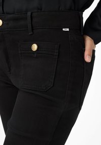 Schwarze Denim-Jeans mit einer Nahaufnahme der Vorderseitentasche, die mit goldenen Beschlägen und einem genähten Etikett am Bund versehen ist. Glatte Textur.