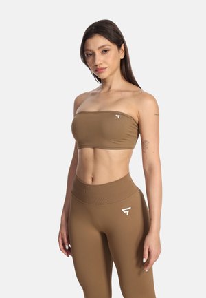 Frau trägt braunes trägerloses Croptop und passende hoch taillierte Leggings mit Logos, steht vor einfarbig hellem Hintergrund.