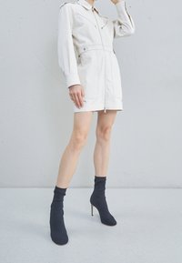 Bílý zip-up šaty s dlouhými rukávy, přiléhavým pasem a předními kapsami. Kombinovány s černými podpatekmi s punčochovými botami. Hladká textilie.