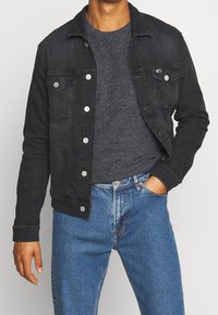 Man som har på sig en svart denimjacka över en mörkgrå prickig t-shirt och blå jeans, med ena handen i fickan.