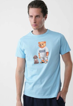 Uomo che indossa una maglietta azzurro chiaro con grafica di un orso con gli occhiali, abbigliamento bianco, sandali, tiene il guinzaglio di un piccolo cane marrone.