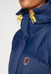 Marineblauwe gewatteerde jas met een hoge kraag, gele trekkoord en een rood-wit logopatch op de borst. Glad, glanzend textiel.