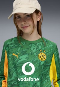 Nuori henkilö, jolla on punaiset hiukset, pukeutuu vihreä-keltaiseen Borussia Dortmundin jalkapallon pelipaitaan ja beigen väriseen Puma-lippikseen harmaata taustaa vasten.