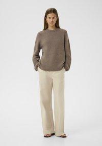 Übergroßer brauner Strickpullover mit Rundhalsausschnitt, kombiniert mit lockeren beige Hosen und flachen schwarzen Sandalen. Schlichtes, strukturiertes Design.